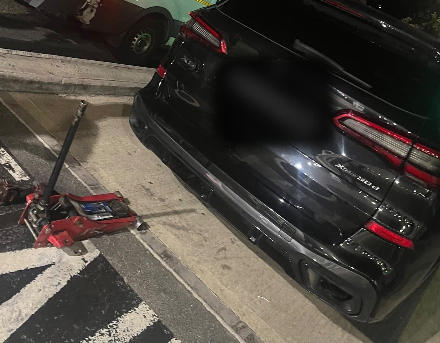 The rear of a black BMW X5 at night with a trolley jack on the pavement beside it.